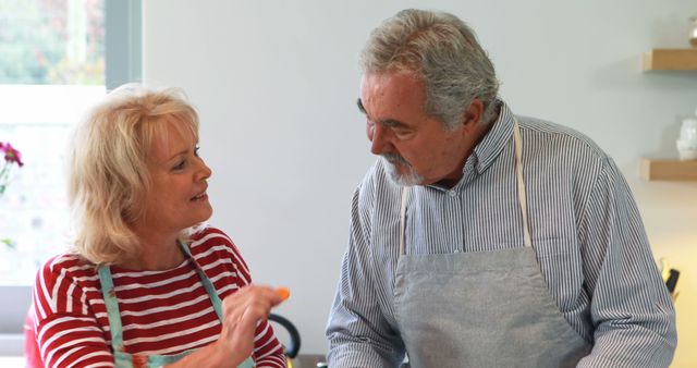 Senior couple preparing food together in a modern kitchen. They are wearing aprons and engaged in a lively conversation, showcasing teamwork and enjoyment. Suitable for lifestyle, family bonding, elderly activities, and healthy living concepts.