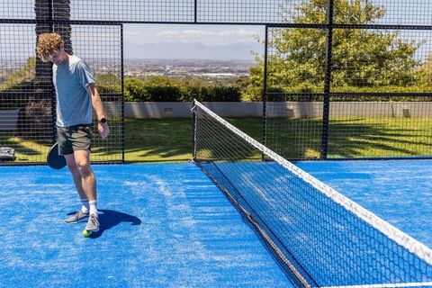 Young man balancing pickleball on foot on vibrant blue outdoor court with city panorama