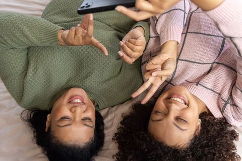 Young women lying on bed taking selfie with peace signs cozy knit sweaters smiling