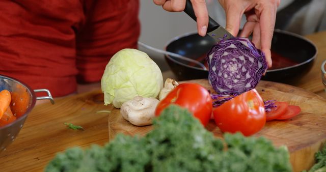 Colorful image depicting person preparing fresh ingredients for a meal. Highlights include a purple cabbage being cut on a wooden board surrounded by mushrooms, tomatoes, and leafy greens. Useful for articles or blogs on healthy eating, recipe ideas, or kitchen activities.