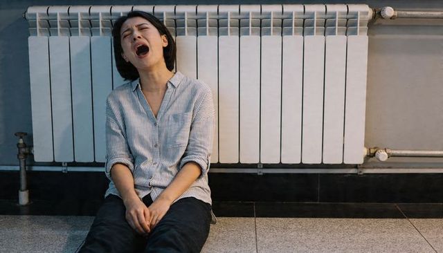 Woman yawning while sitting on tiled floor by radiator, clasping hands, tired corridor mood