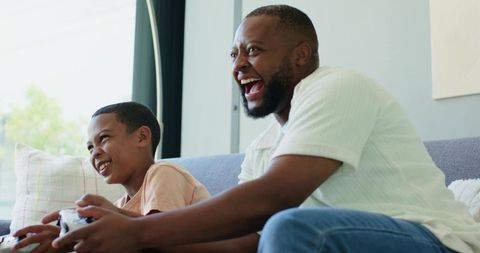 African American Father and Son Laughing Playing Video Games on Couch in Bright Room