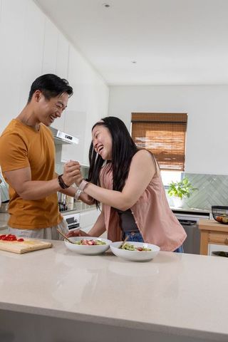 Asian couple laughing while preparing salad at bright modern kitchen island