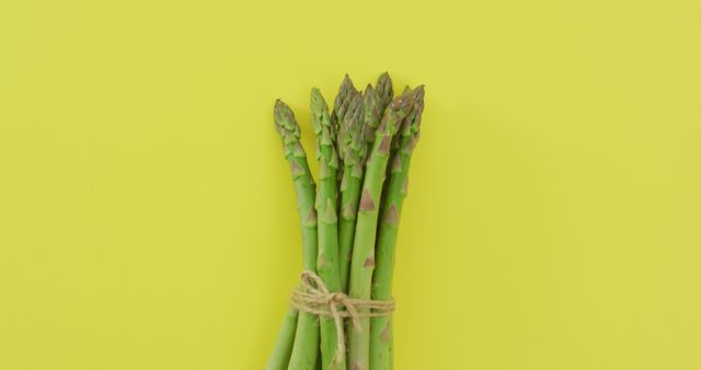 Bunch of fresh green asparagus stalks tied with a string against a vibrant yellow background, showcasing a clean and minimalistic look. This image is perfect for use in healthy lifestyle blogs, vegan recipe illustrations, diet and nutrition articles, or organic product promotions.
