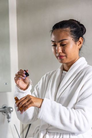Asian woman applying facial serum in white bathrobe at minimalist bathroom sink