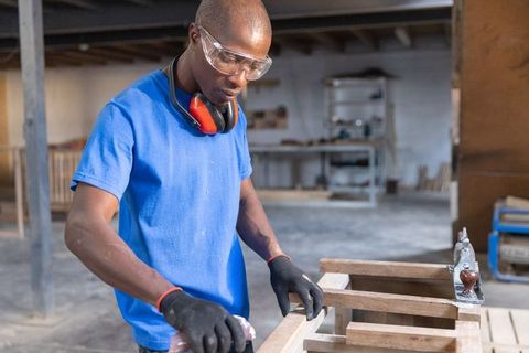 African american craftsman sanding wooden frame wearing safety gear in workshop