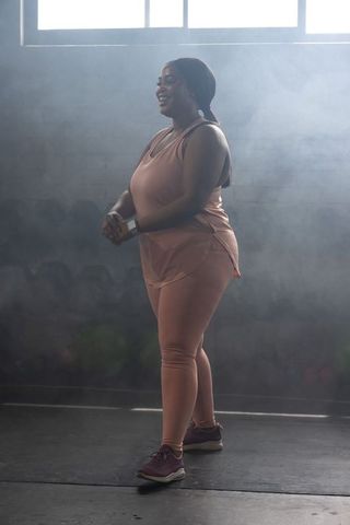Smiling african american woman standing in sunlit gym wearing rose workout set and sneakers