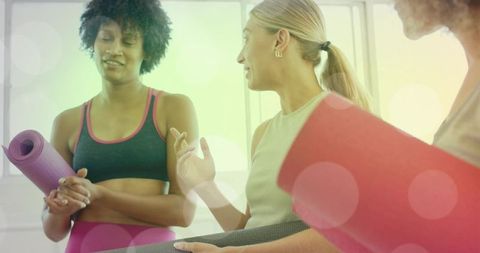Diverse women holding yoga mats chatting and preparing for class in bright sunlit studio