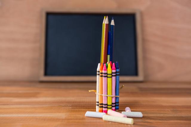 Colorful crayons and pencils arranged on a wooden table with a chalkboard in the background. Ideal for educational content, back-to-school promotions, art and craft projects, and creative learning resources.