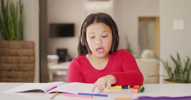Girl wearing headphones, focusing on homework at home. Ideal for use in education blogs, remote learning resources, child development articles, and online tutoring promotions. Conveying themes of concentration, home schooling, and the importance of a comfortable study environment.