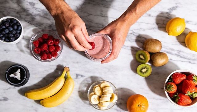 Hands adding raspberry to pink berry smoothie on marble countertop with fresh fruit