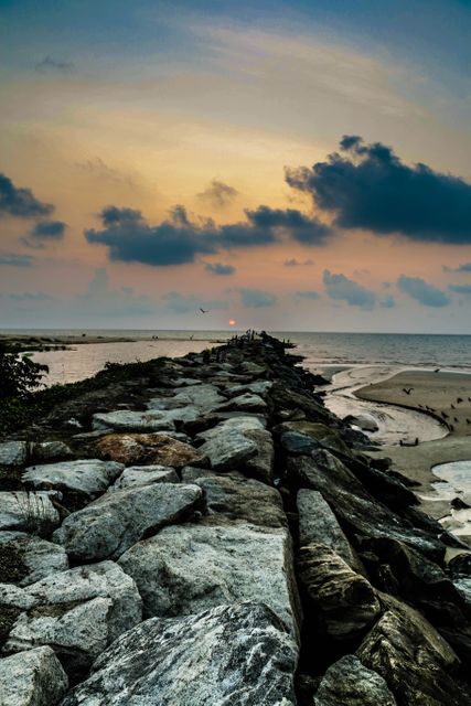 Rocky pier stretches into ocean under dramatic evening sky with vibrant sunset hues, dark clouds adding contrast. Ideal for travel blogs, nature-themed websites, or background for relaxation content. Perfect for social media posts, inspirational quotes, and vacation advertisements.