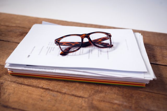 Eyeglasses resting on a stack of documents on a wooden office desk. Ideal for illustrating themes of work, business, organization, and professional environments. Useful for articles, blogs, and presentations related to office life, productivity, and workplace organization.