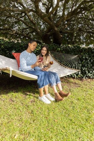 Diverse friends relaxing on hammock sharing smartphone and drink in sunny garden