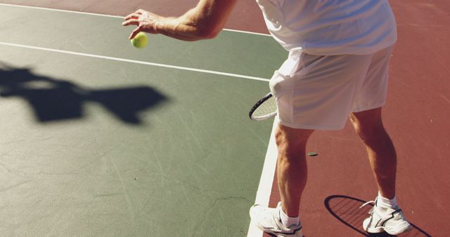 Close-up of tennis player wearing white outfit serving on outdoor tennis court during sunny day. Focus is on the ball and lower body of player. Useful for sports, fitness, training, or recreation concepts. Can be used in articles, advertisements, blogs focusing on tennis, athletic performance, or healthy lifestyle.