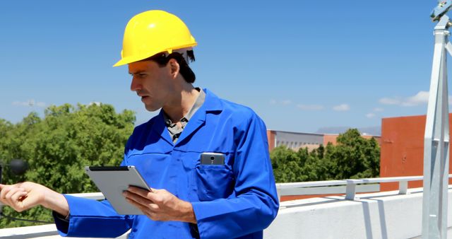 Young Caucasian man in blue workwear inspects a construction site. He's focused on safety and project management, tablet in hand for efficiency.