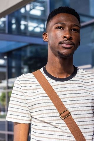 African man standing looking up at modern glass building wearing striped tee and tan strap