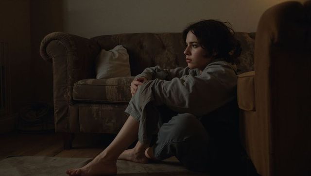 Contemplative woman sitting on floor hugging knees beside worn sofa in warm low light
