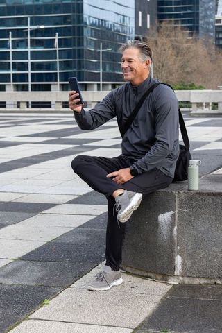 Active mature man sitting in city plaza checking phone wearing activewear with water bottle