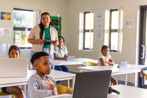 Elementary teacher assisting diverse students with laptops and tablets in contemporary classroom