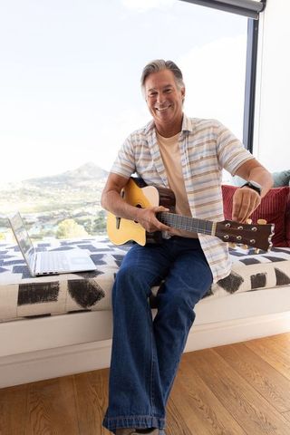 Mature man tuning acoustic guitar on sunny window bench with laptop and scenic view