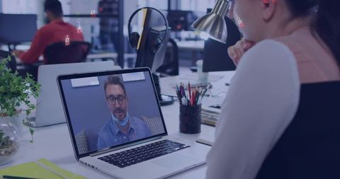 Woman watching remote video meeting on laptop at open-plan coworking desk with headset