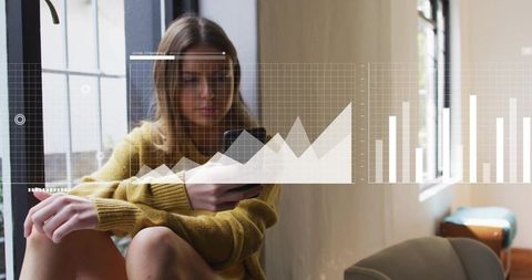 Young woman sitting holding smartphone with digital charts overlay in sunlit lounge