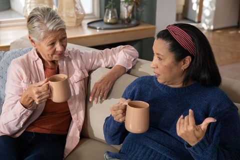 Diverse senior friends holding matching mugs and chatting on cozy beige sofa in sunlit living room