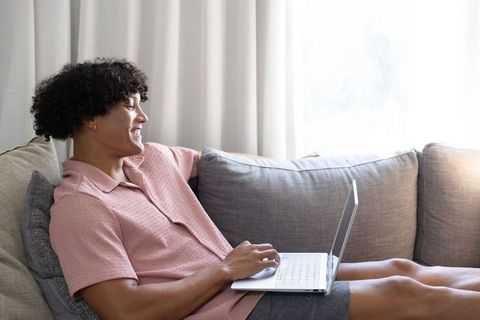 Relaxed young man using laptop on light-gray couch in bright minimalist living room