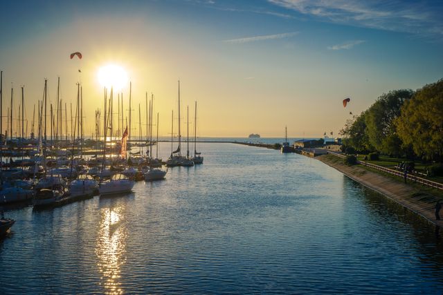 Beautiful harbor scene at sunrise with sailboats docked. Gentle morning light casts a serene atmosphere over calm waters with people strolling near the lush green park. Perfect for travel blogs, nature photography, or tourism promotions highlighting serene coastal destinations.