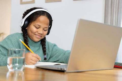 Young girl studying from home wearing headphones, smiling while writing homework at laptop
