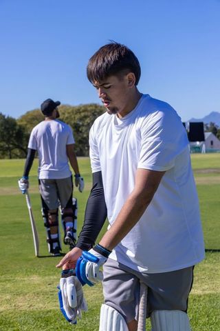 Young cricketer adjusting gloves on sunny practice field while teammate walks with bat