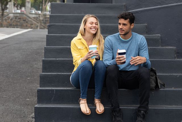 Happy, fashionable Caucasian couple out and about in the city during the day, siting on steps, talking and smiling, bothe holding takeaway coffees.