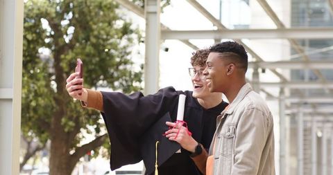 Graduating friends taking selfie on urban campus celebrating diploma and mortarboard