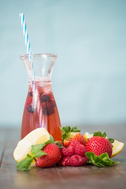 Berry cocktail in a glass bottle with a striped straw, surrounded by fresh strawberries, raspberries, lemon slices, and mint leaves on a wooden surface. Ideal for themes related to summer refreshments, healthy beverages, fruit garnishes, or non-alcoholic drink recipes.