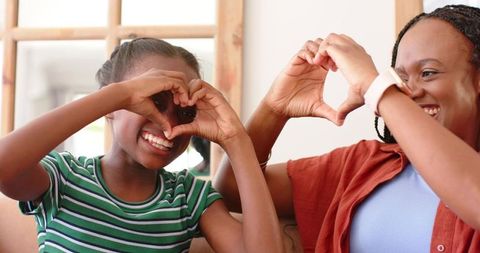 African american mother and daughter making heart hands while laughing on cozy sofa