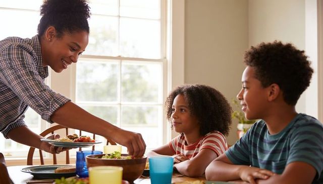 Smiling mother serving warm family dinner at sunlit wooden table with kids