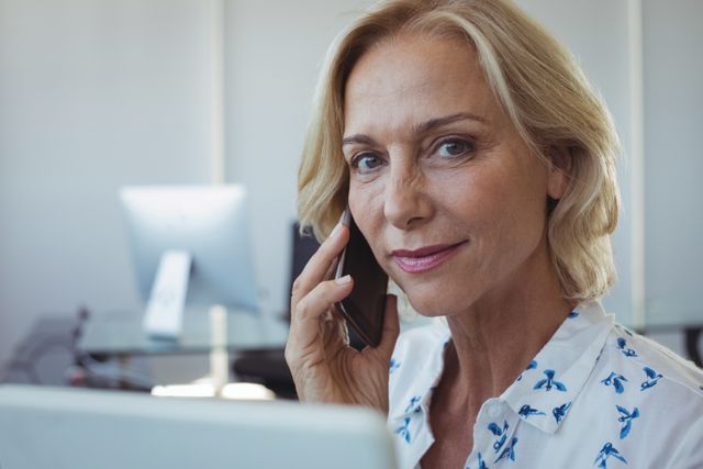 Mature businesswoman talking on mobile phone in modern office. Ideal for use in business, corporate communication, technology, and professional career-related content. Perfect for illustrating concepts of leadership, confidence, and workplace communication.