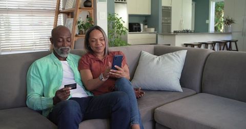 Couple using smartphone and credit card on sofa making online payment in modern lounge