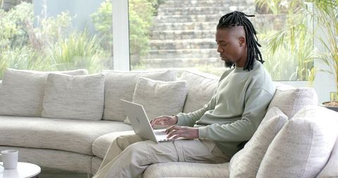 African American man working remotely on laptop in bright living room with garden view