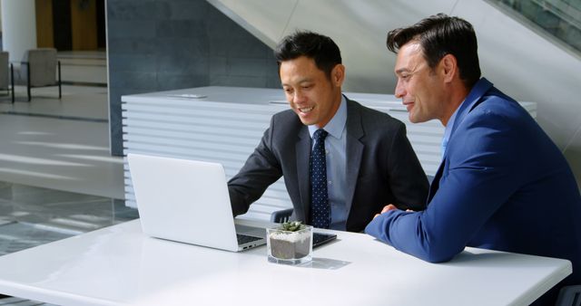 Two businessmen in suits are sitting at a white table, actively discussing a work project using a laptop in a modern office environment with a plant centerpiece. This can be used for depicting teamwork, business meetings, strategy planning, and professional workplace interactions.