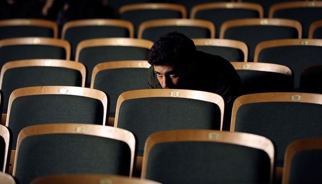 Solitary man leaning over theater seat number 15 in empty auditorium, moody cinematic lighting