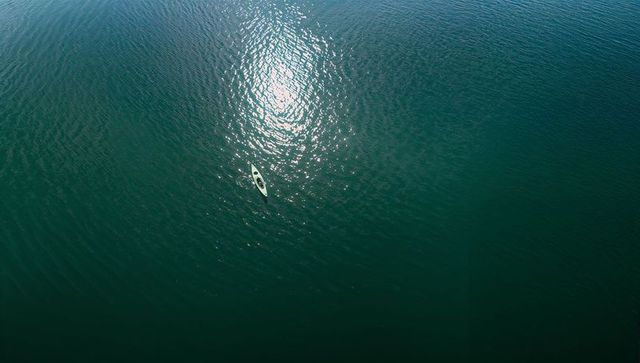Solitary kayaker paddling across sunlit teal open water aerial minimalist seascape