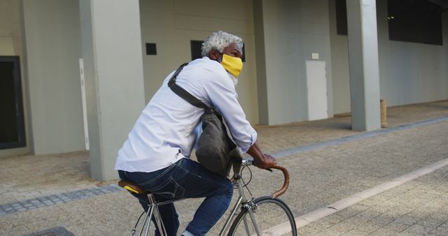 African american senior man wearing face mask riding bicycle in corporate park. hygiene and social distancing during coronavirus covid-19 pandemic.