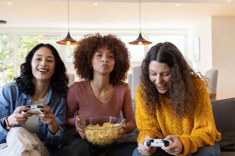 Diverse women playing video games and snacking on chips on sofa in sunlit living room