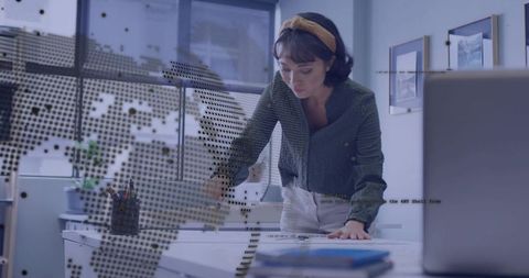 Creative designer leaning over desk drafting sketches with laptop and world map overlay