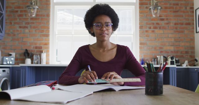 Woman is sitting at a kitchen table, surrounded by books and notes, holding a pen and ruler while studying. She appears focused on her work with glasses on and an afro hairstyle. The setting includes a brick wall and blue accents, with ample natural light from a window. Perfect for illustrating educational themes, homework, remote study environment, or a work from home scenario.