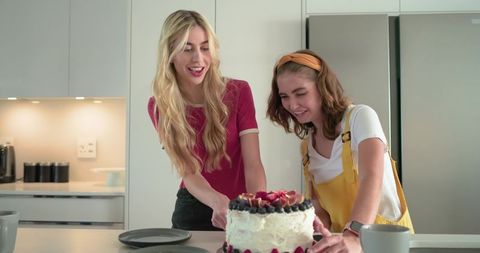 Two friends decorating frosted berry cake at kitchen island, joyful home baking moment