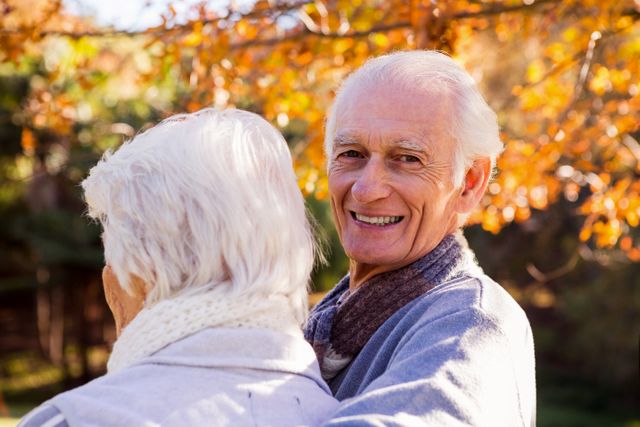 Senior couple enjoying a beautiful autumn day in the park. The man is smiling warmly while his wife stands in the foreground. Perfect for use in advertisements, brochures, or articles related to senior living, retirement, health and wellness, and outdoor activities for the elderly.