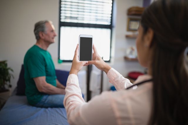 Therapist using mobile phone to photograph senior male patient in hospital ward. Useful for illustrating modern healthcare practices, patient documentation, elderly care, and the use of technology in medical settings.